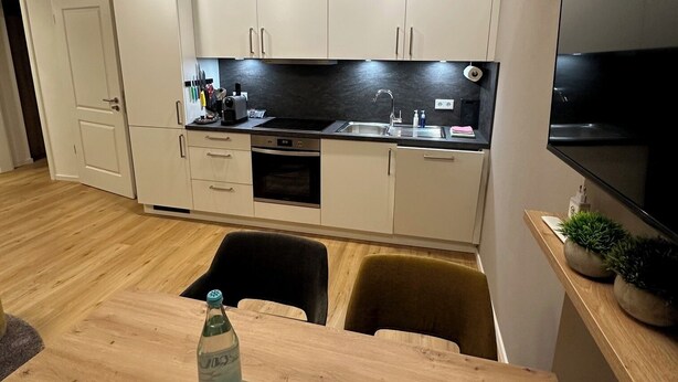 A modern kitchen with white cabinets, a built-in oven, and a black countertop. There is a dining table with chairs and a bottle of water on it. - Apartment Ahrblick (1-4 persons) | Top vacation apartment with a view of the Ahr (Altenahr)