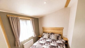 Une chambre à coucher avec une tête de lit en bois, un lit recouvert d’une literie à motifs, une table de chevet, une fenêtre avec des rideaux et un radiateur.