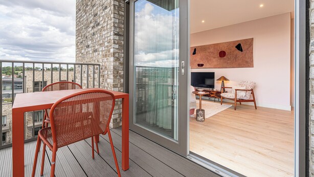 Un balcon avec une table et des chaises, une porte-fenêtre coulissante donnant sur un espace intérieur équipé d’un téléviseur, d’un tapis et d’un fauteuil.