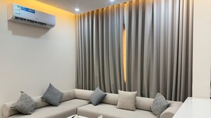 A modern living room with a sectional sofa, two white coffee tables, and a large window with vertical blinds. - Aswar Plus for serviced apartments (Tabuk)