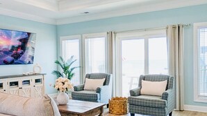 Ein Wohnzimmer mit einem beigen Sofa, zwei Sesseln, einem Couchtisch und einem Deckenventilator. Es gibt Fenster mit Vorhängen, ein Bild an der Wand und eine Vase mit Blumen auf einem Schrank.