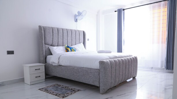 Une chambre à coucher moderne avec un lit recouvert d’un tissu gris, du linge de lit blanc, une petite table de chevet et un ventilateur fixé au mur.