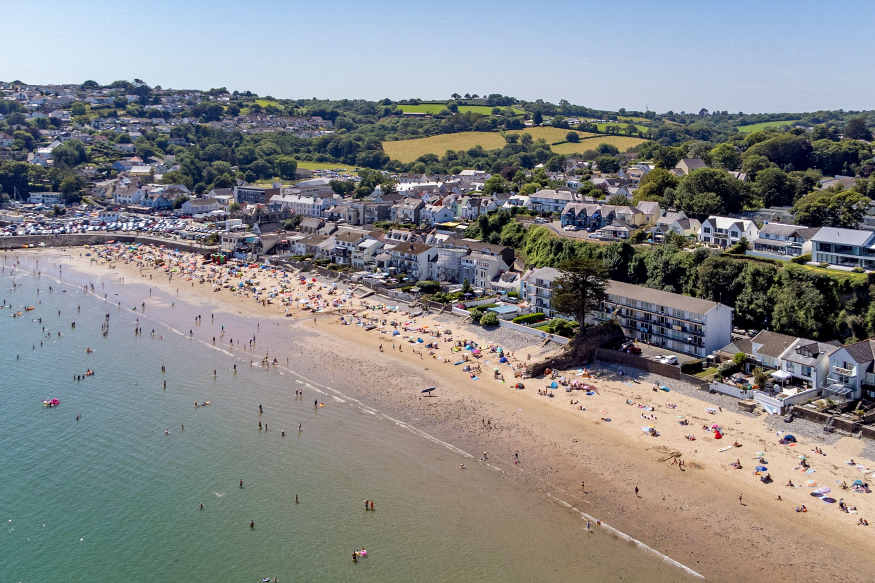 Luftaufnahme einer Küstenstadt mit Sandstrand, Wohnhäusern und einer hügeligen Landschaft im Hintergrund.