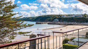 Une vue sur la plage depuis un balcon doté d’une balustrade, un ciel dégagé et une côte lointaine parsemée de bâtiments.