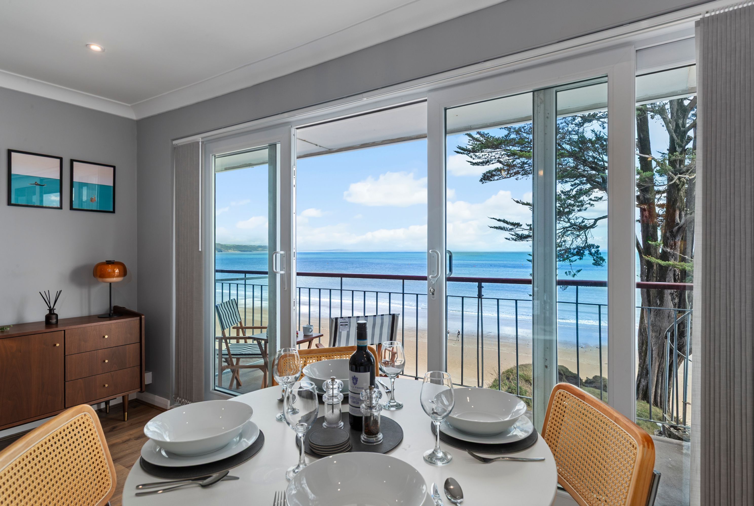 A dining area with a table set for four, overlooking a beach with a clear view of the ocean and a balcony with a railing.