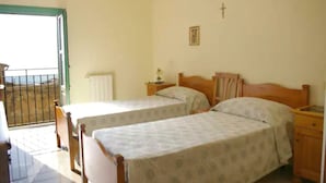 Quarto com duas camas de solteiro, uma pequena mesa de cabeceira com abajur, cabeceira de madeira, janela com vista e uma cruz na parede.