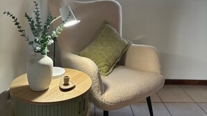 Een gezellige kamer met een ronde houten tafel, een witte vaas met groene planten, een groen kussen op een beige fauteuil en uitzicht op het omliggende landschap door een raam.