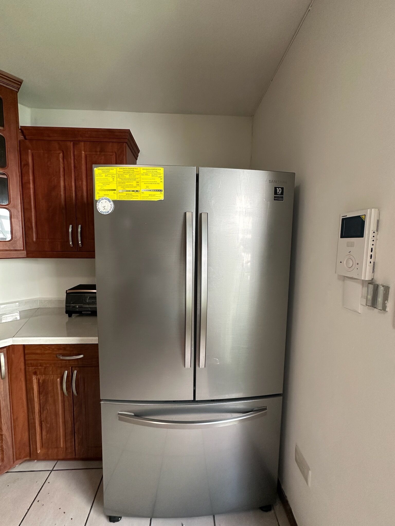 Un refrigerador de acero inoxidable con pantalla digital, situado en una cocina con armarios de madera y encimera blanca.
