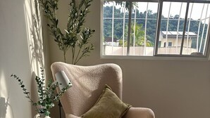 Een gezellige kamer met een comfortabele fauteuil, een klein bijzettafeltje met een vaas en een kopje, een geweven vloerkleed en een raam met uitzicht op groen.