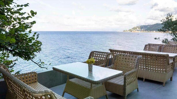 An outdoor seating area with wicker furniture and a glass table, overlooking a body of water. - Venesia Hotel (Arsin)