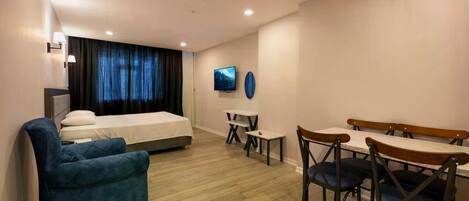 A hotel room with a bed, a blue armchair, a dining table with chairs, a TV, and a small table with a chair.