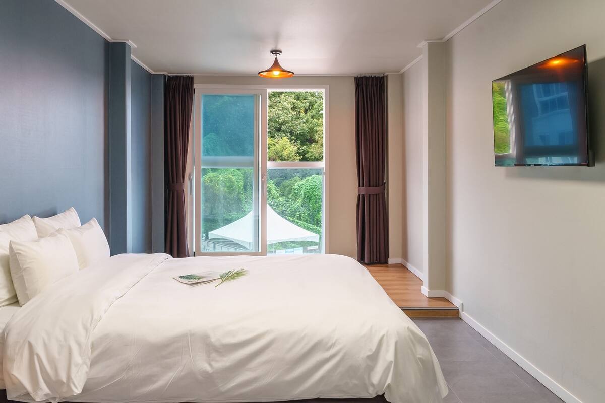 Una cama bien hecha con sábanas y almohadas blancas, una lámpara de techo, un ventanal con cortinas y un televisor de pantalla plana montado en la pared.