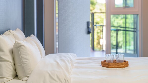 Een bed met witte lakens en kussens, een rieten dienblad met glazen en een uitzicht op groen door een raam.