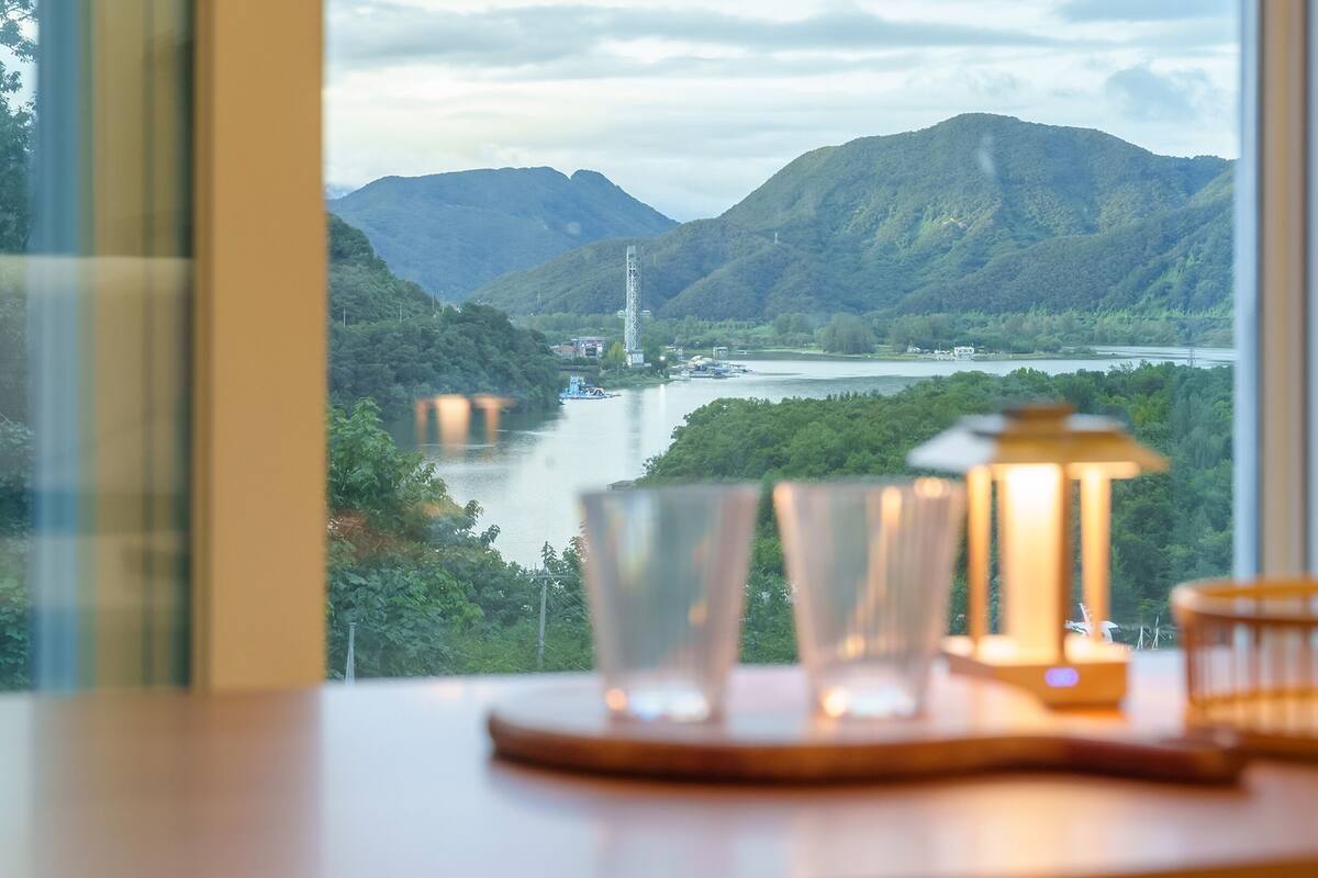 Vista de un río con montañas al fondo, una mesa con vasos y una vela, y un marco de ventana.