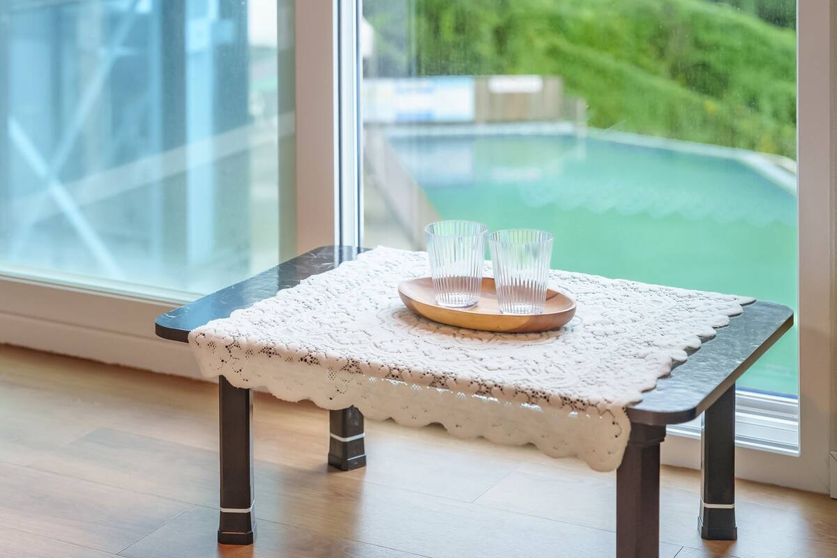 Una pequeña mesa de madera con mantel de encaje y dos vasos transparentes sobre una bandeja, ubicada junto a una ventana con vistas a un paisaje verde.