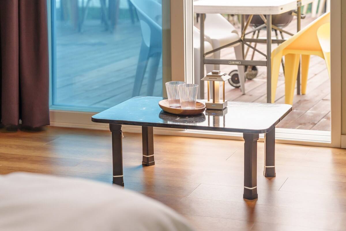 Una pequeña mesa con bandeja y vasos, una ventana con cortinas, y vistas a muebles de exterior.