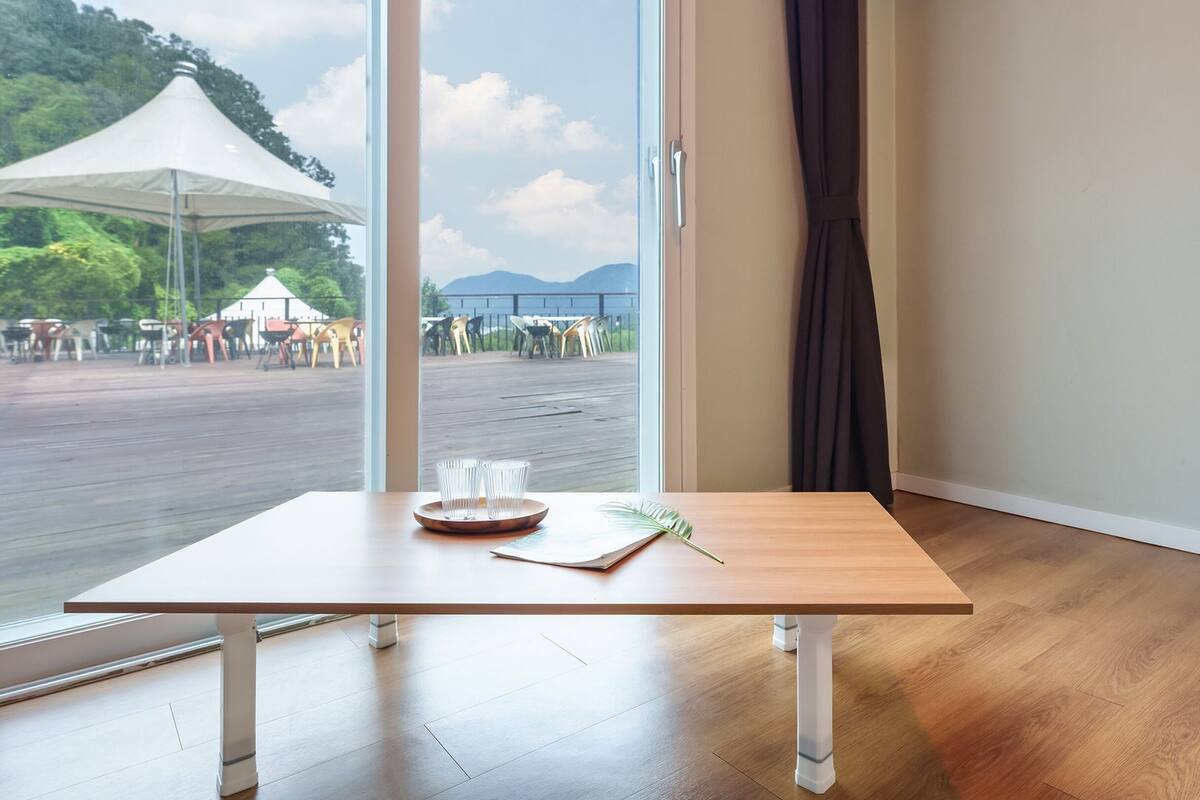 Una habitación con una mesa de madera, un ventanal con vistas a un área de estar exterior con sillas, y una cortina en la ventana.