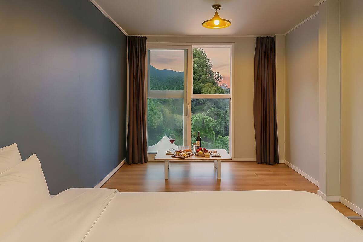Un dormitorio con una cama grande, una mesa con comida y bebida, y vista a la vegetación por una ventana.