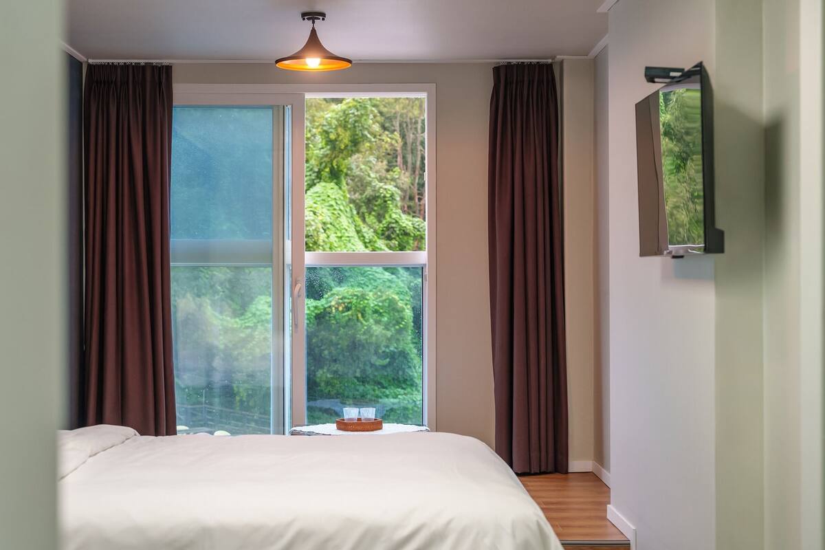 Un dormitorio con cama, ventana con cortinas, lámpara de techo y televisor montado en la pared.