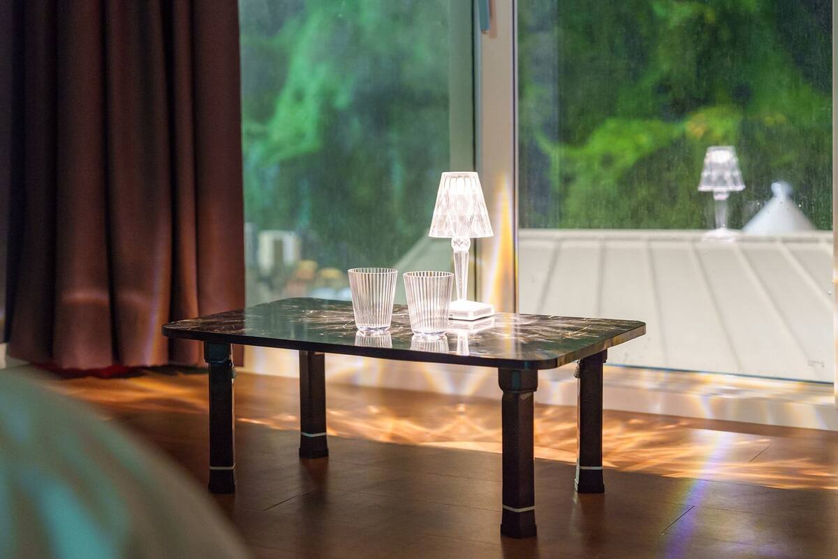 Una pequeña mesa con una lámpara y dos vasos sobre un piso de madera, cerca de una ventana con cortinas.