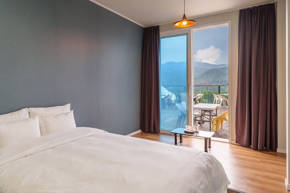 Habitación de hotel con cama, una mesita pequeña con una silla y vistas a un balcón con mobiliario exterior.