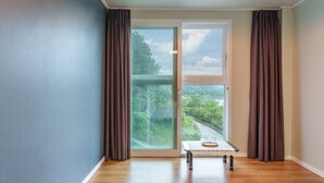 Ein Zimmer mit einer Schiebetür zu einem Balkon, einem kleinen Tischchen mit Tablett sowie Blick auf eine Straße und Grünflächen.