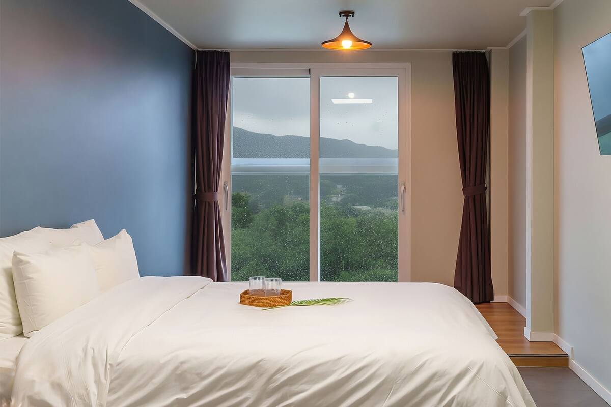 Un dormitorio con una cama, un televisor montado en la pared y un amplio ventanal con cortinas.