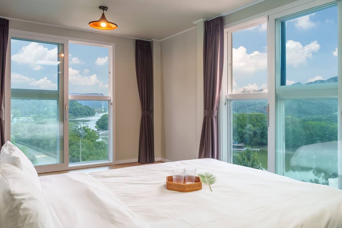 Habitación de hotel con una cama grande, dos ventanas con cortinas, vista a un río y montañas, y una mesita con vasos y un mantel.