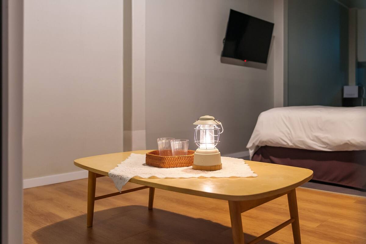 Una habitación con una mesa de madera, una lámpara blanca y una cama con cubre cama blanco.