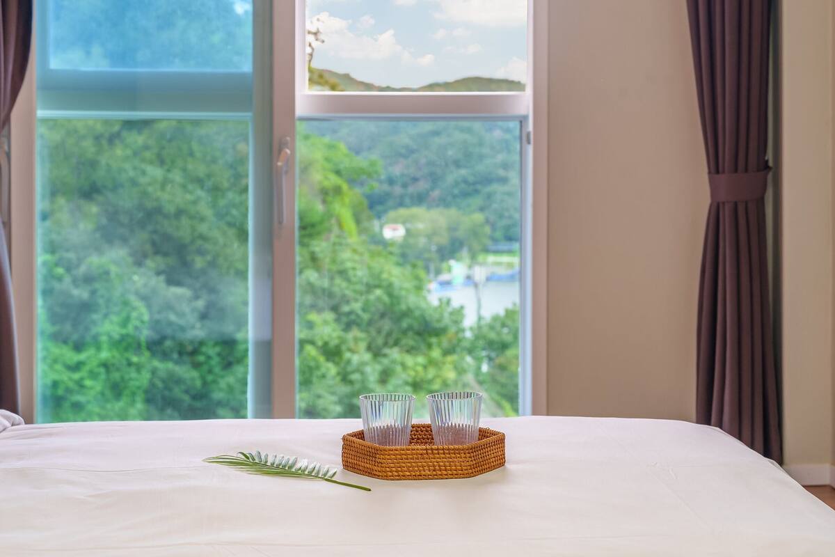 Habitación de hotel con una cama, vistas a zonas verdes y un lago, y una cesta con vasos.
