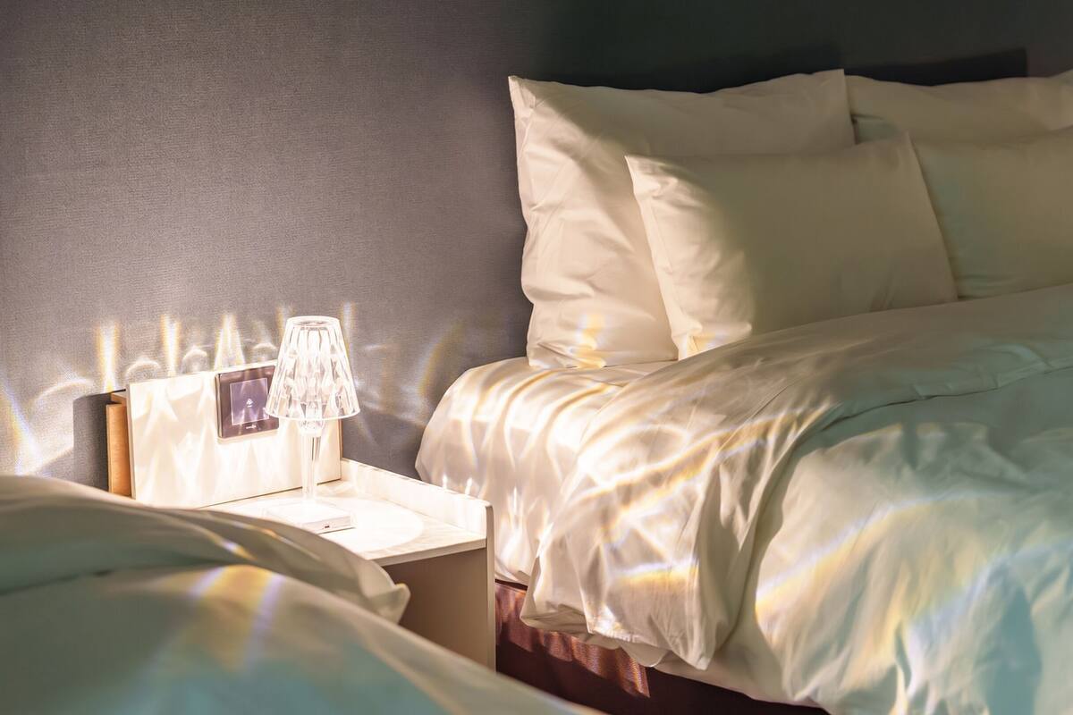 Una cama bien hecha con sábanas y almohadas blancas, una mesita de noche con una lámpara, y un reloj de pared.