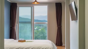 A bedroom with a bed, a window with curtains, a view of greenery, and a ceiling light.