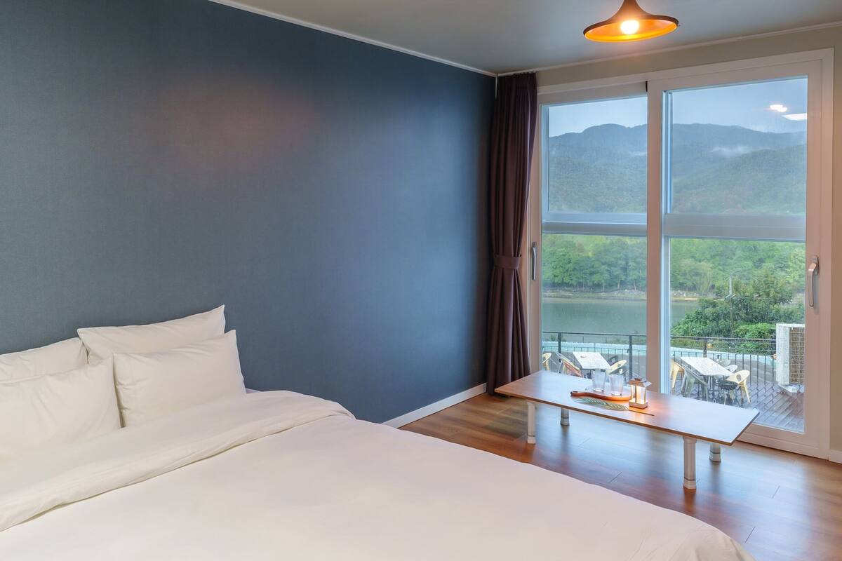 Un dormitorio con una cama grande, una mesita pequeña y vistas a un lago y montañas a través de un ventanal amplio.