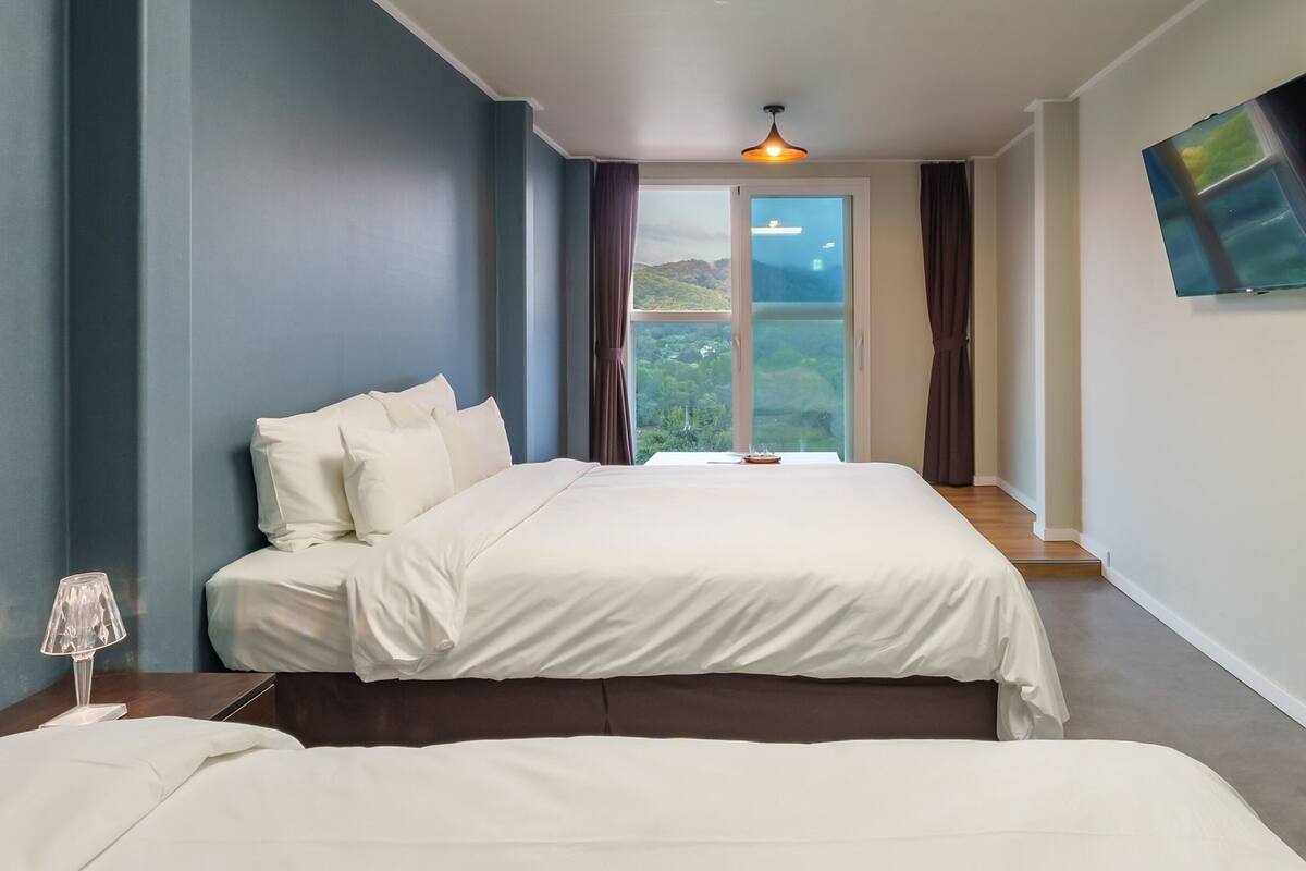 Habitación de hotel con una cama grande, una cama más pequeña, una mesita de noche, una lámpara y vistas al exterior a través de un ventanal amplio.