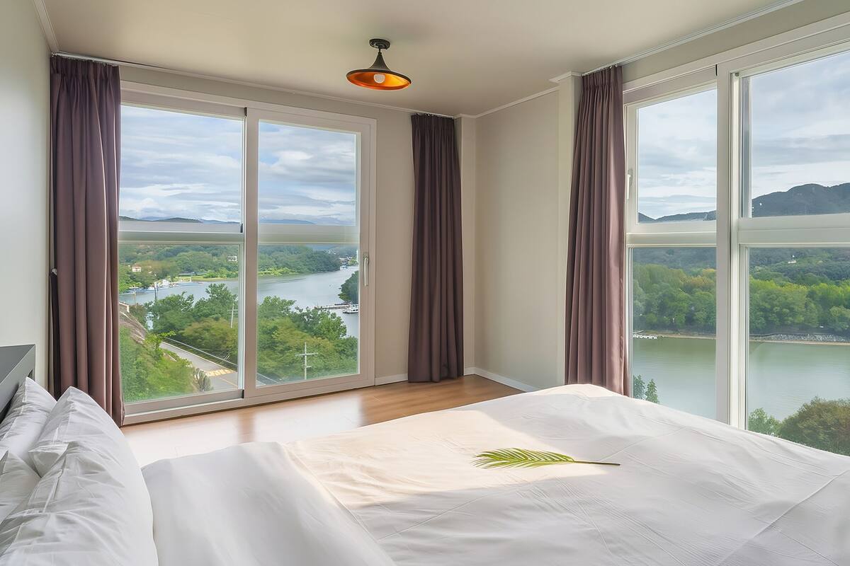 Habitación de hotel con una cama grande, vistas a un río y montañas, y una lámpara en el techo.