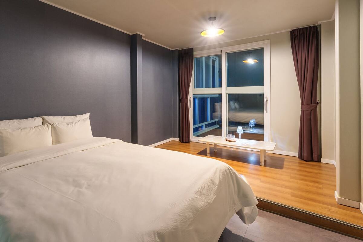 Un dormitorio con una cama grande, una ventana con cortinas, una mesita con lámpara y vistas a un balcón.