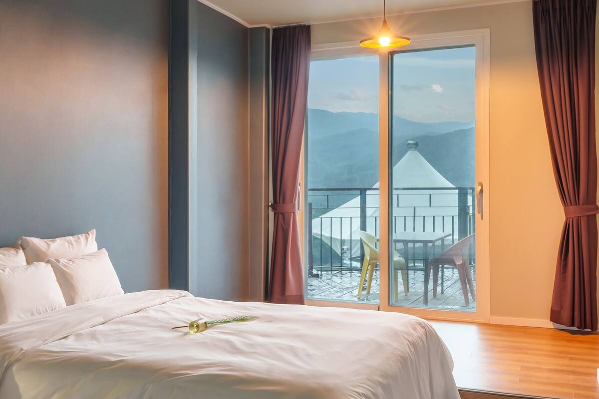 Un dormitorio con una cama grande, vistas a un balcón con mobiliario exterior y montañas al fondo.
