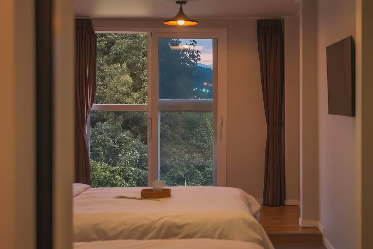 Un dormitorio con un ventanal grande, una cama, una mesita de noche y una lámpara colgante.