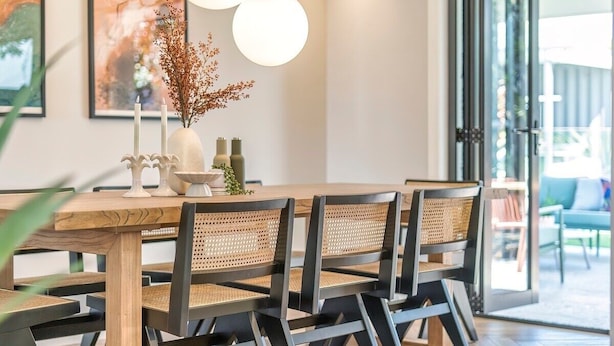 Un comedor con una mesa de madera y sillas negras con asientos trenzados, un jarrón con ramas secas y dos luces esféricas fijadas a la pared.
