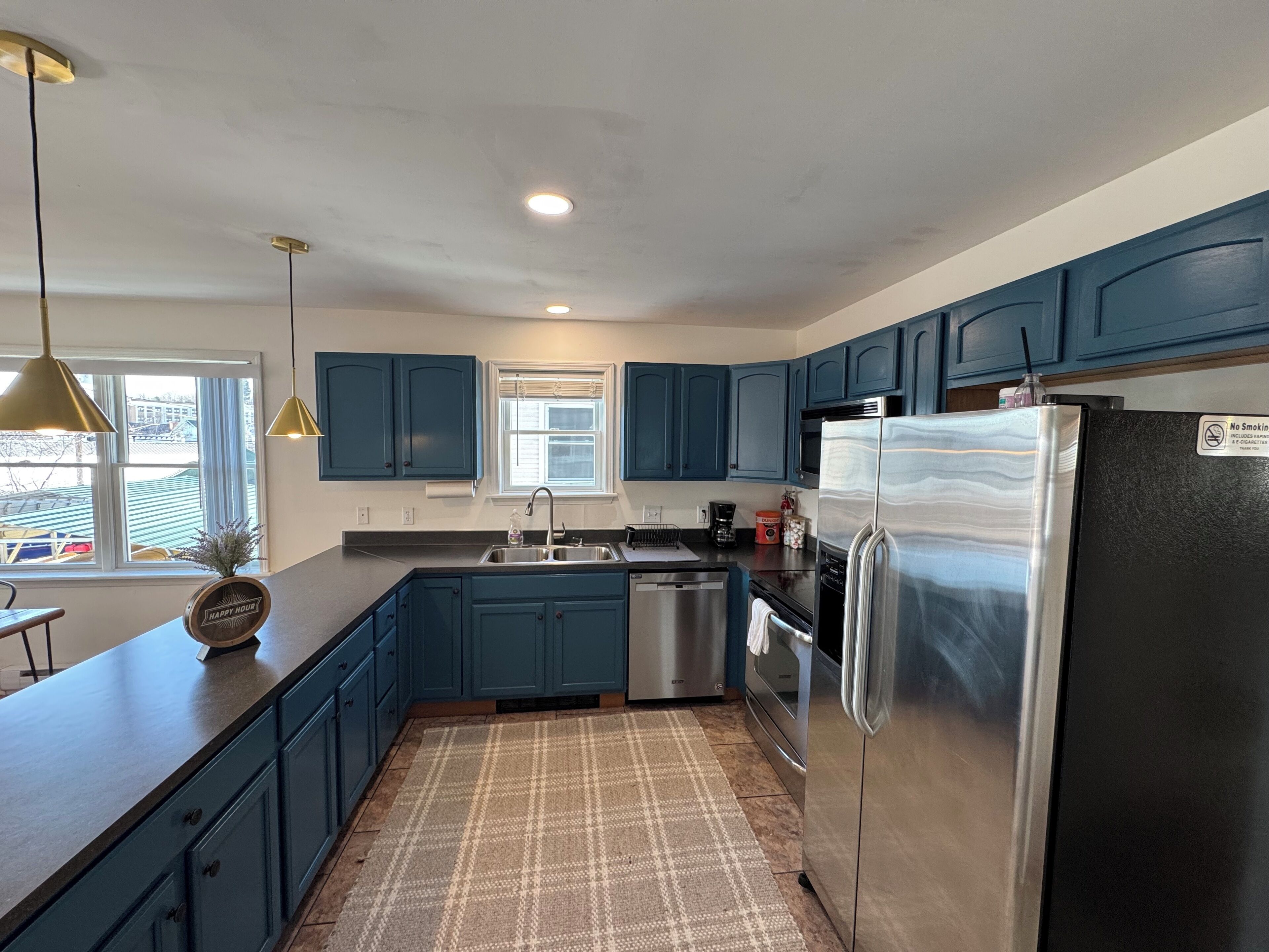 A modern kitchen with blue cabinets, stainless steel appliances, a large window, and a patterned rug.