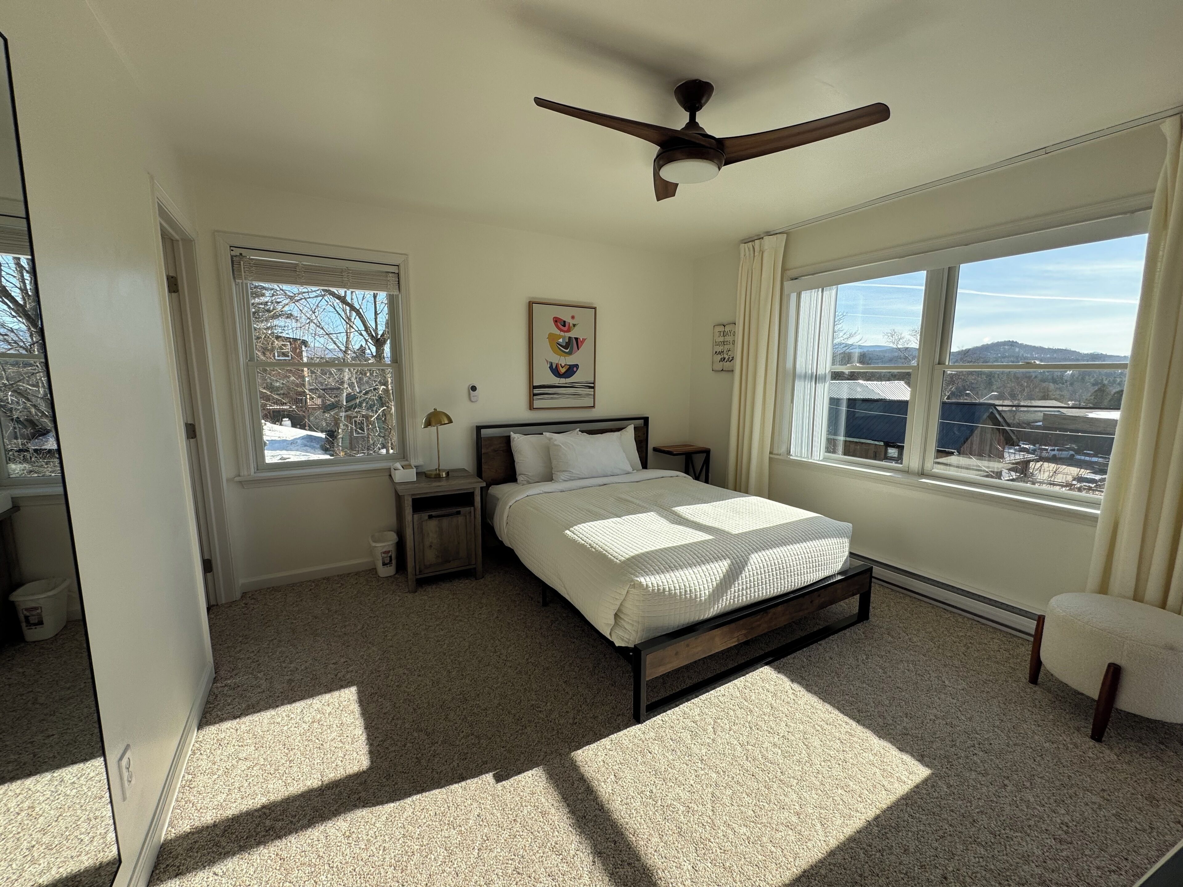 A bedroom with a bed, a nightstand, a window with a view of the outside, and a ceiling fan.