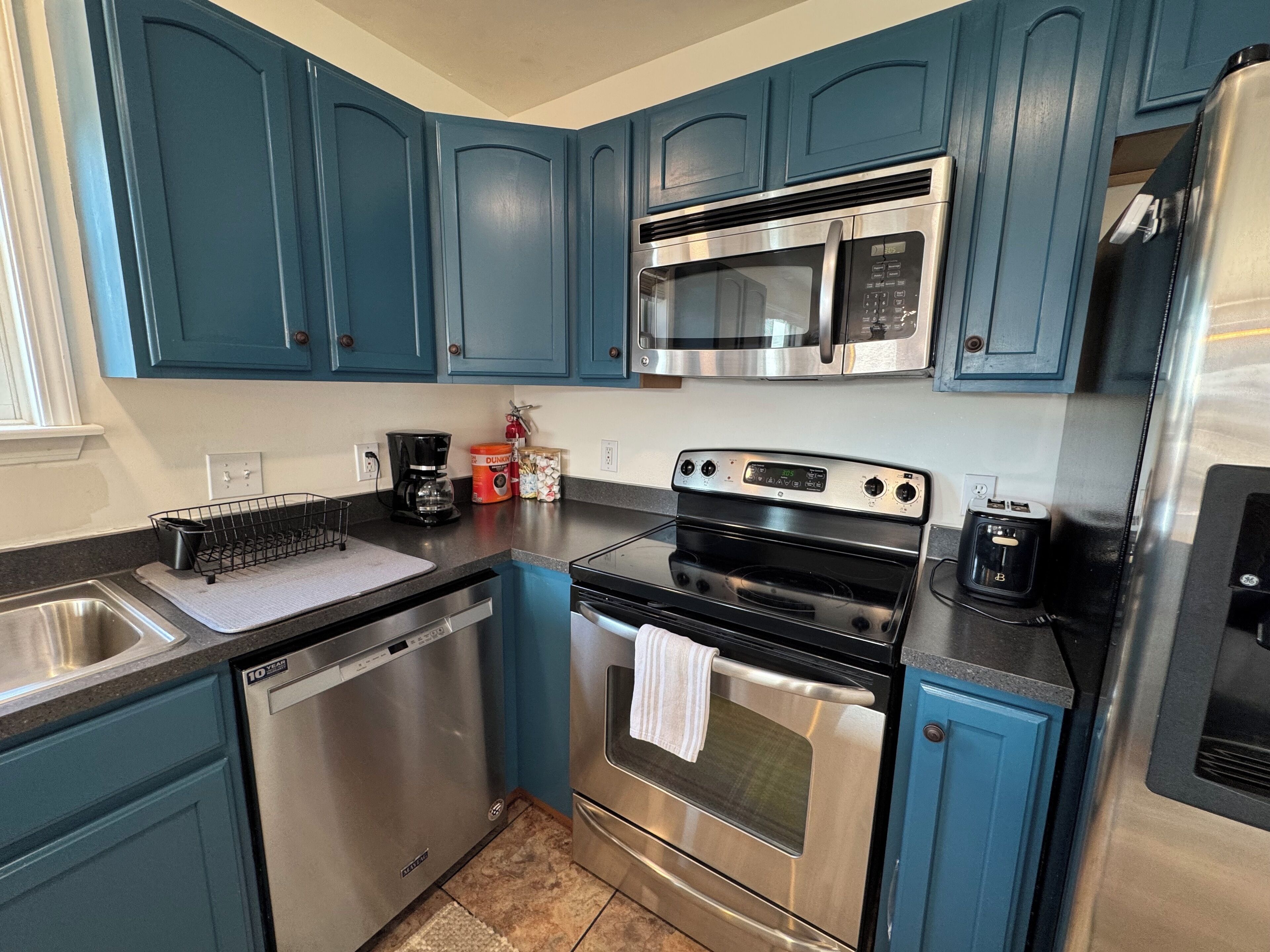 A modern kitchen with teal cabinets, stainless steel appliances including a microwave, oven, and dishwasher, and a coffee maker on the counter.