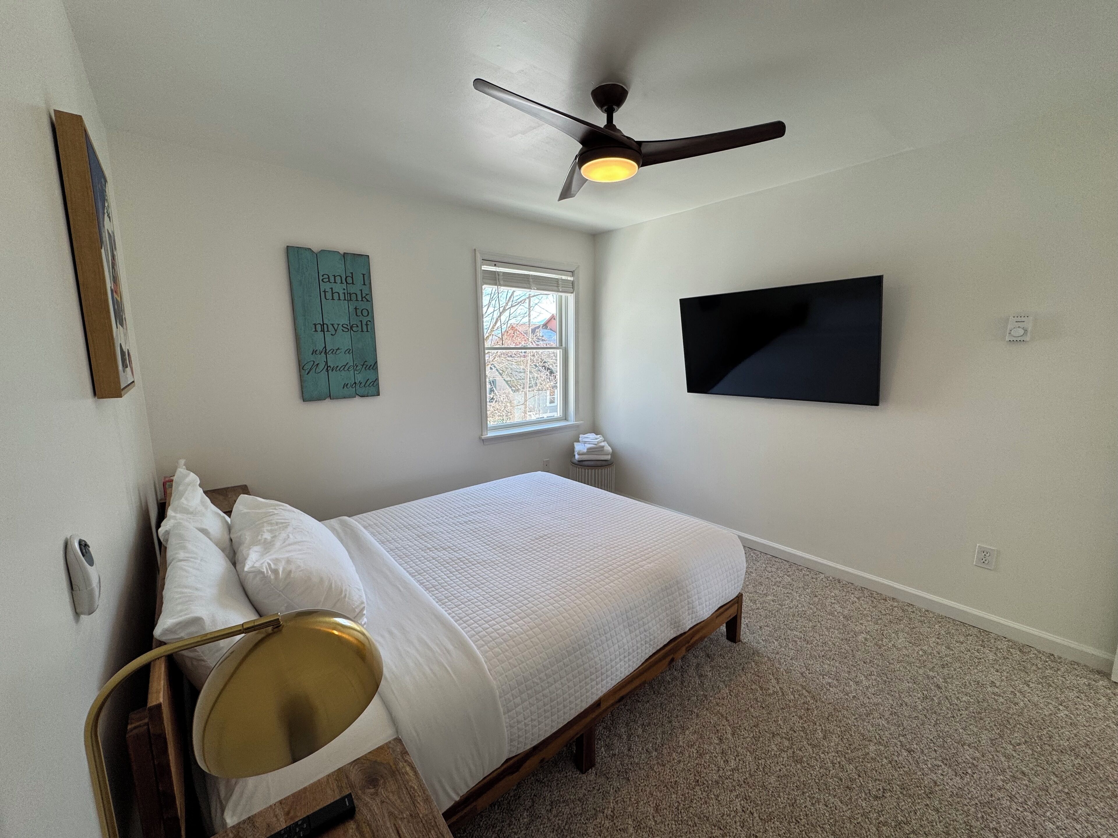Une chambre à coucher avec un lit, un ventilateur de plafond, un téléviseur fixé au mur, une fenêtre et une citation inspirante sur le mur.