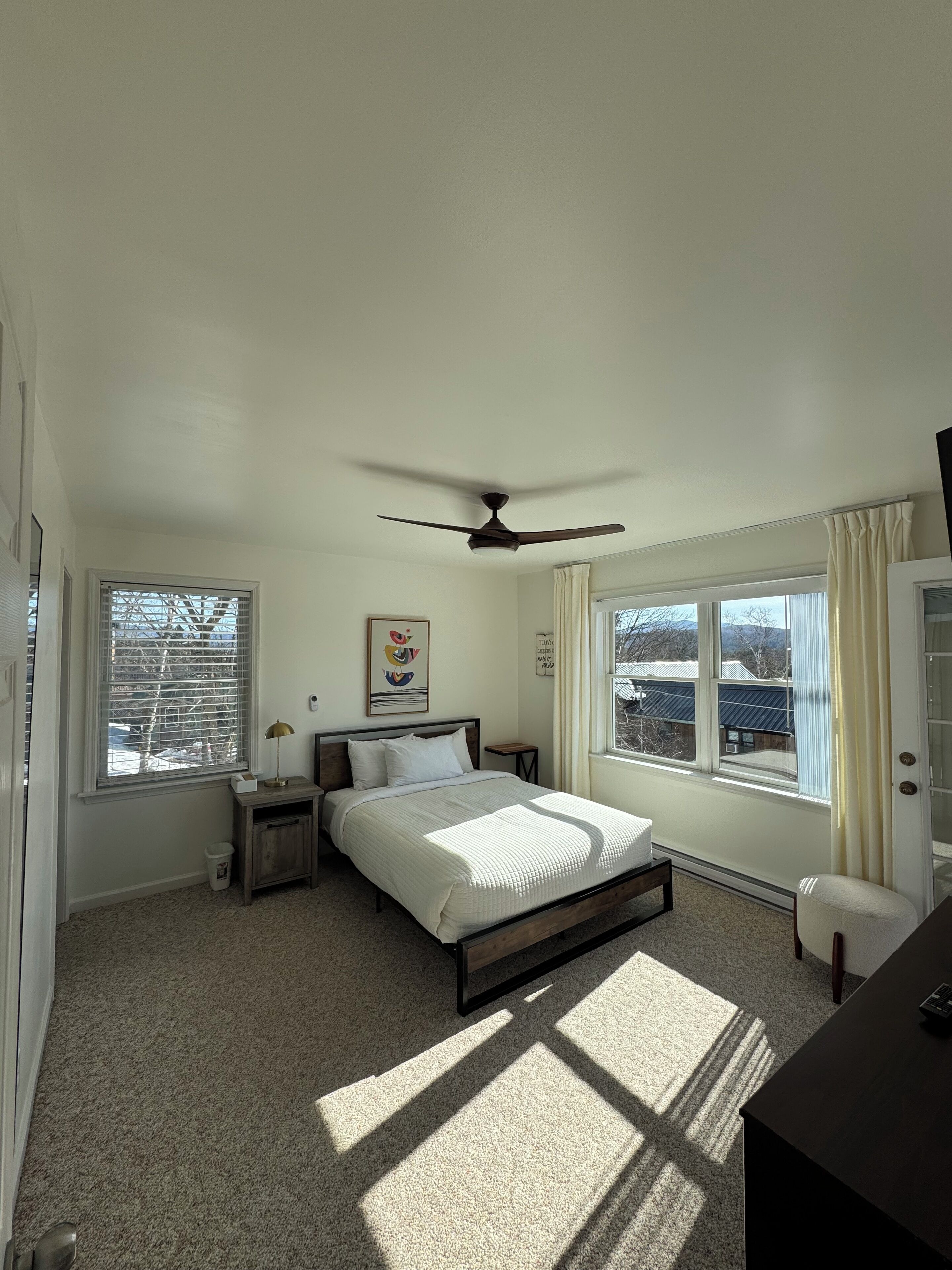 A bedroom with a bed, a ceiling fan, a bedside table, a nightstand, a window with blinds, and a view of the outside.