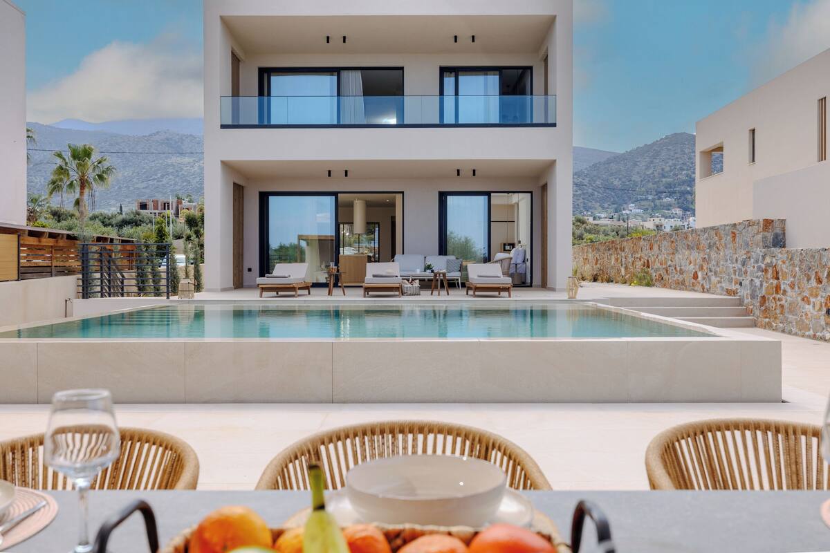 A modern two-story building with a swimming pool, outdoor seating, and a view of mountains.