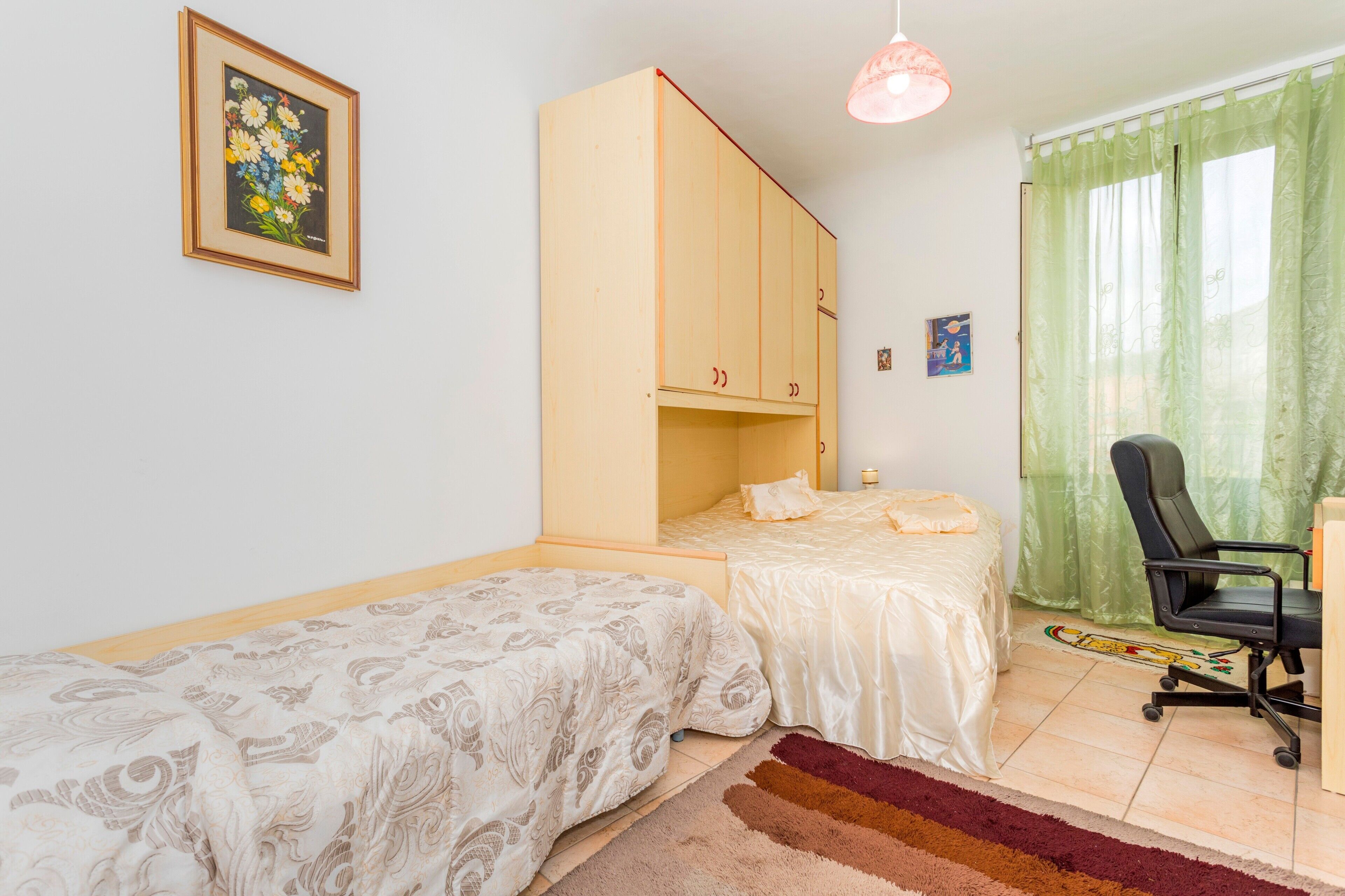 A room with two beds, a wardrobe, a desk with a chair, a hanging light, and a painting on the wall.