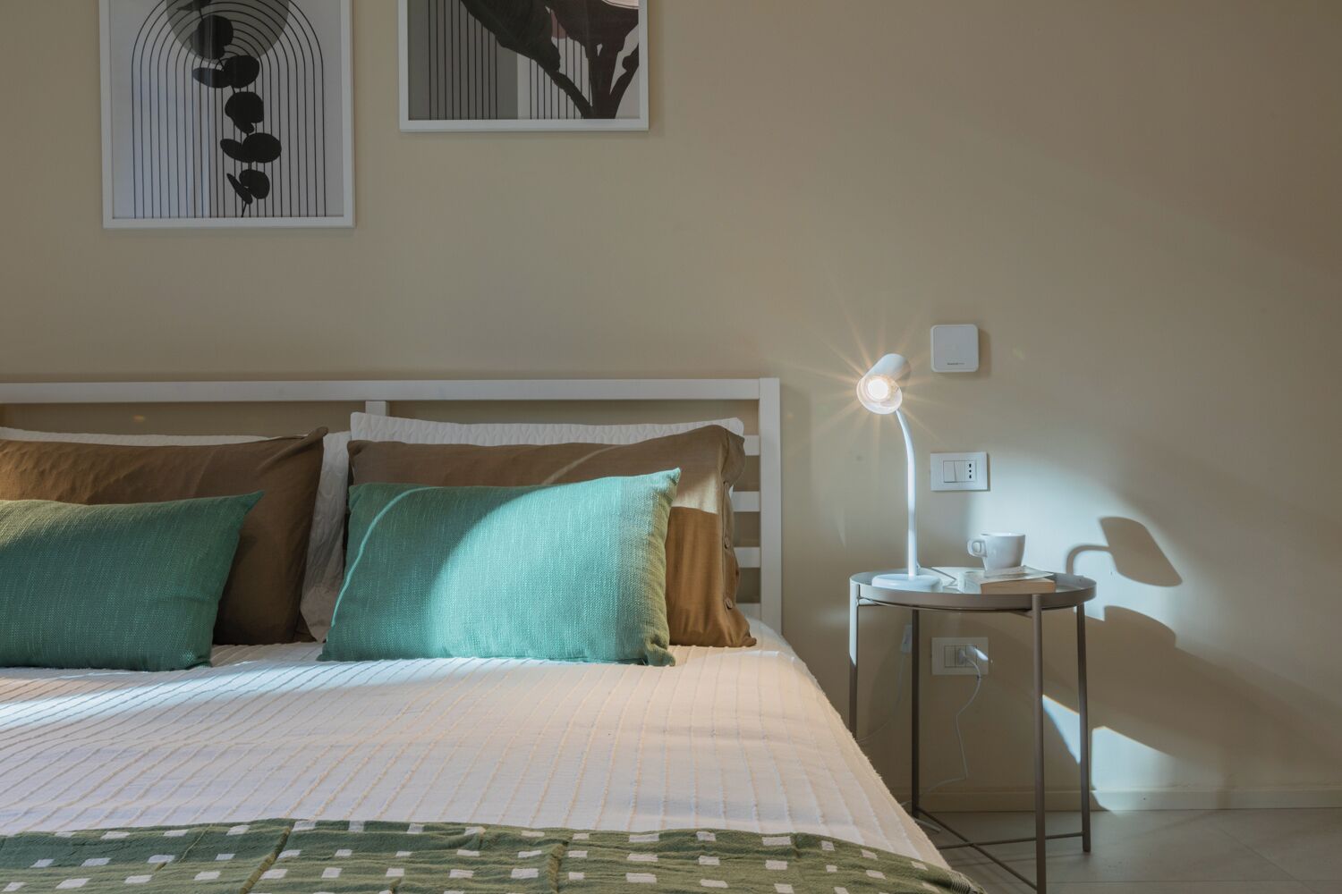 A neatly made bed with a white headboard, two pillows, and a green throw blanket. There is a bedside table with a lamp, a cup, and a small plant. Two framed artworks are hung on the wall above the bed.