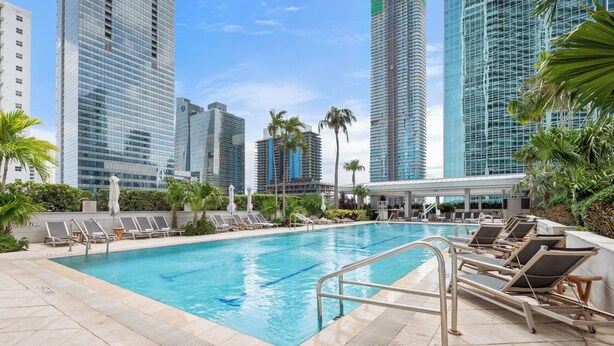 Una piscina con sillones de descanso, rodeada de edificios altos y palmeras.