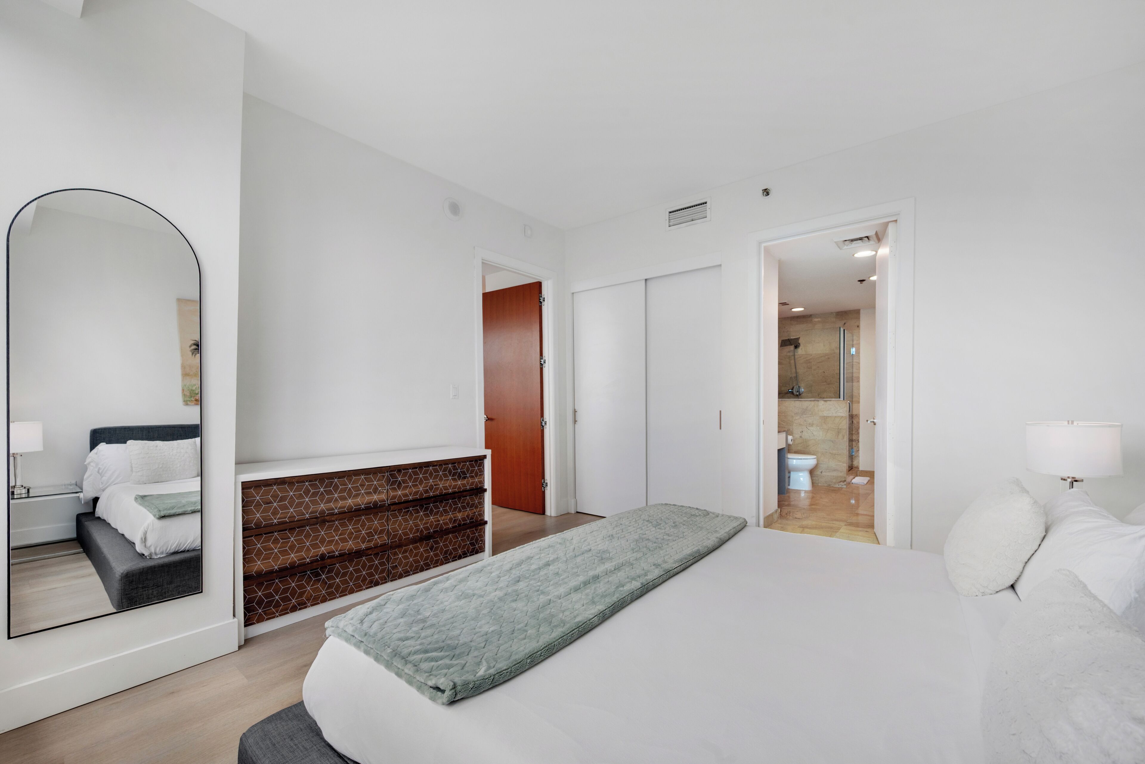 A modern hotel room with a bed, a large mirror, a wooden dresser, and a bathroom visible through an open door.