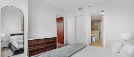A modern hotel room with a bed, a large mirror, a wooden dresser, and a bathroom visible through an open door.